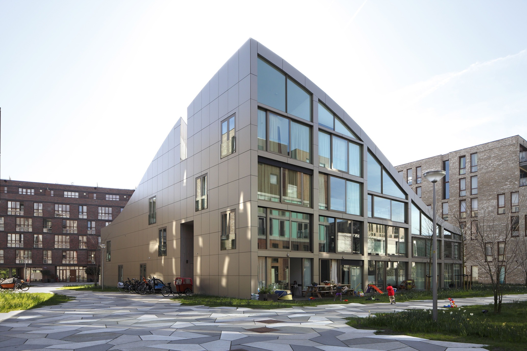 Park-facing facade of Blok K / Verdana with large windows, ground-level outdoor space, and a sloping roofline.