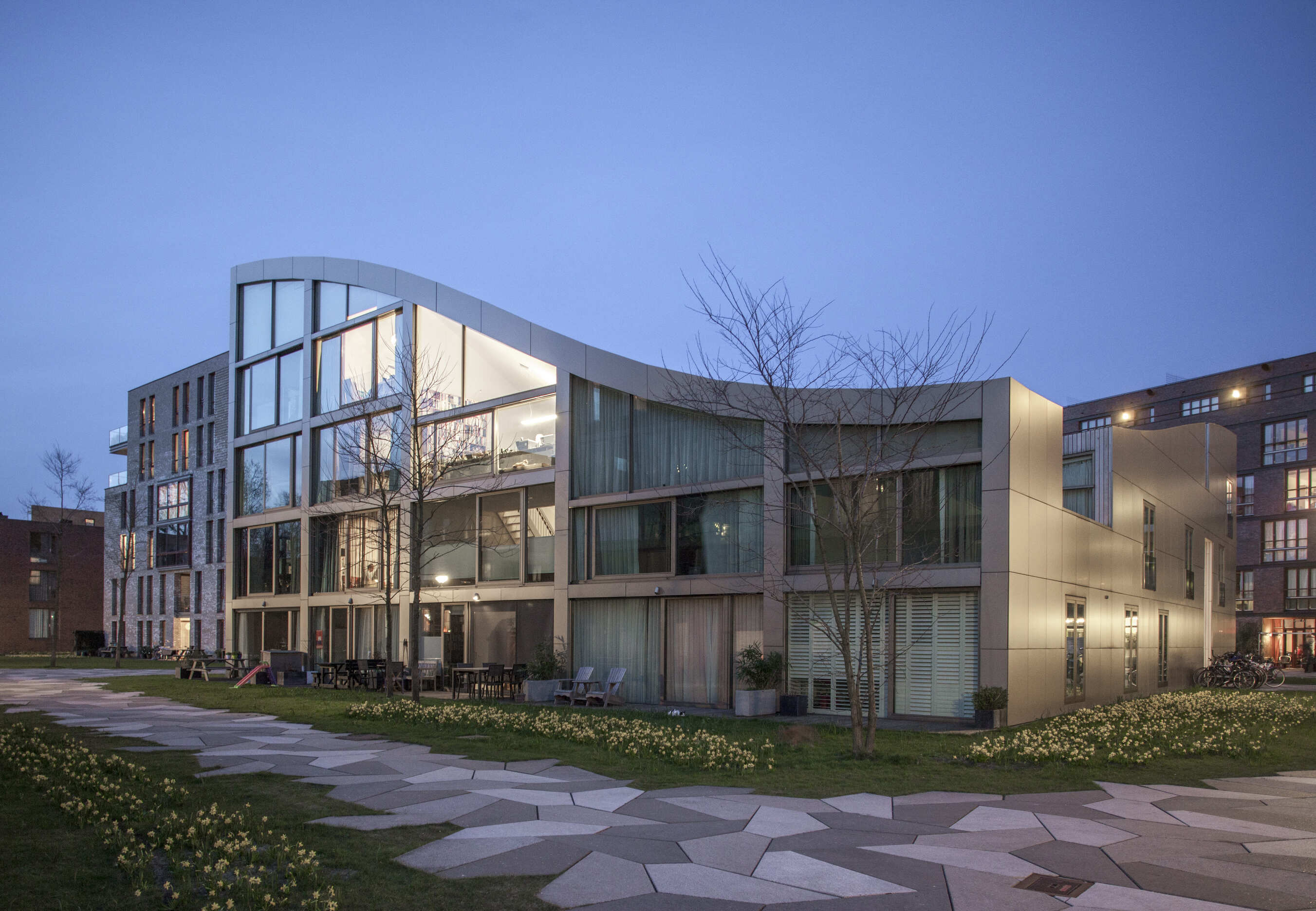 Evening view of Blok K / Verdana from the park, with illuminated interiors and the roofline visible above the facade.
